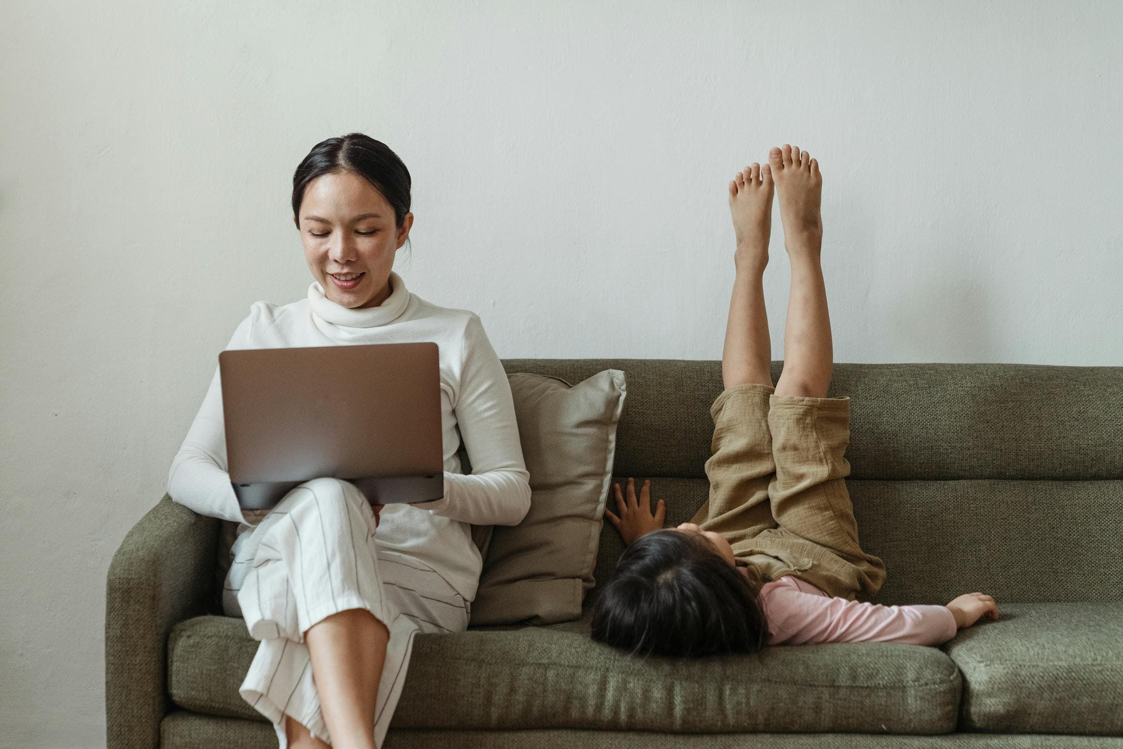 Mother on laptop while daughter plays beside her on the couch